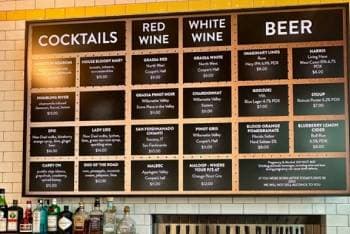 Drink menu board above a self-serve tap with liquor bottles on shelves